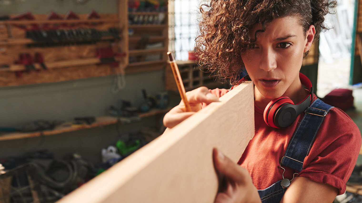 Young concentrated female carpenter with curly hair holding wooden plank and estimating its length before sawing Schlagwort(e): woman, female, young, pretty, curly, denim, overall, concentrated, focused, carpenter, carpentry, joinery, timber, wood, wooden, plank, measuring, measurement, length, eye, sight, tool, skill, manual, equipment, professional, craft, craftsman, handyman, occupation, working, woodworking, workshop, lumber, woman, female, young, pretty, curly, denim, overall, concentrated, focused, carpenter, carpentry, joinery, timber, wood, wooden, plank, measuring, measurement, length, eye, sight, tool, skill, manual, equipment, professional, craft, craftsman, handyman, occupation, working, woodworking, workshop, lumber