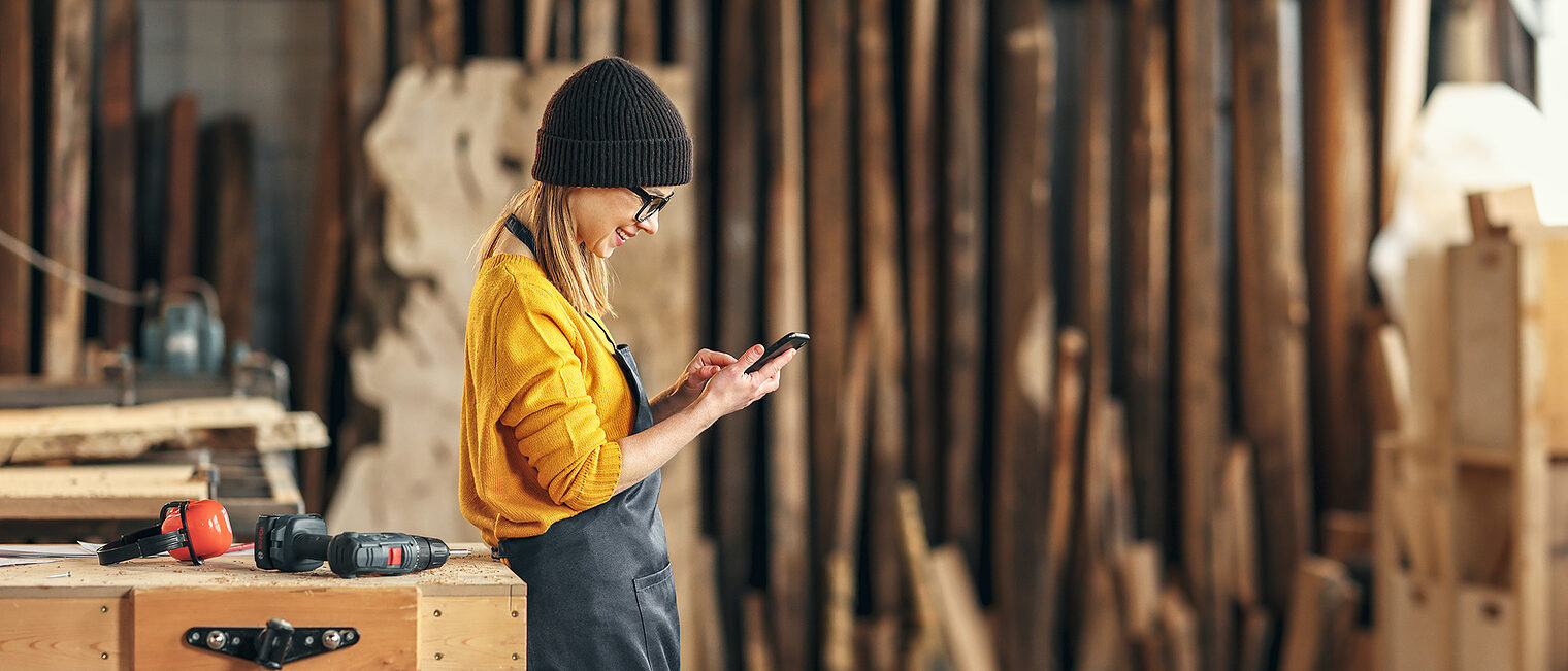 Junge Frau in der Holzwerkstatt mit Smartphone. Bild: JenkoAtaman / stock.adobe.com