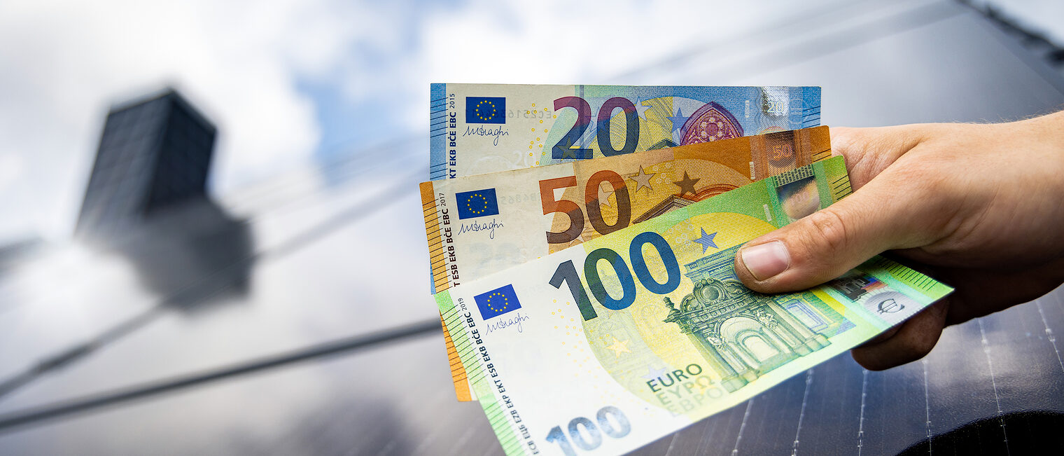 Hand hält Euro-Banknoten, Solarpanel, Photovoltaikanlage. Bild: photoschmidt / stock.adobe.com