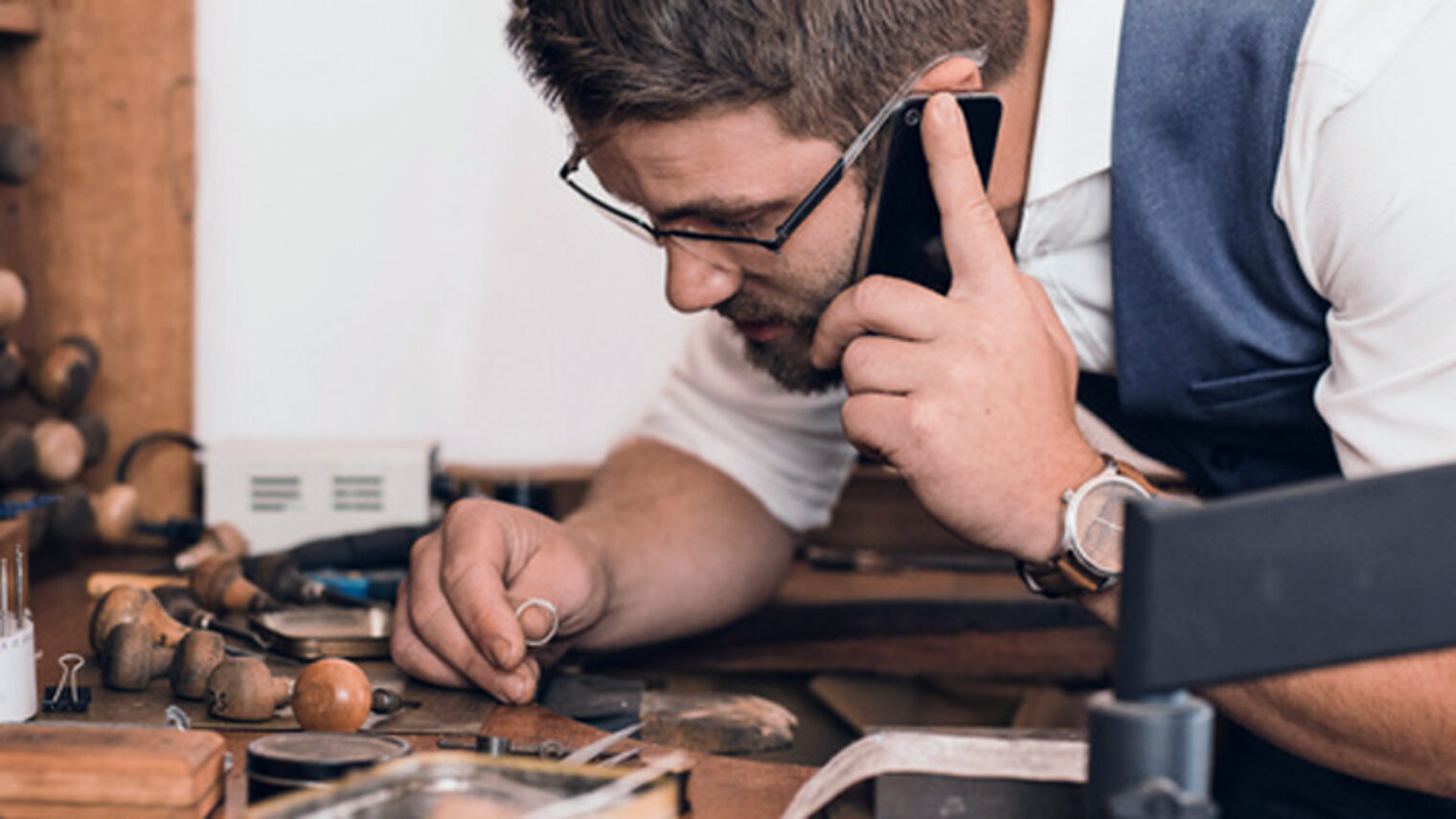 Young jeweler examining a ring and talking on a cellphone while leaning over a workbench full of tools in his shop Schlagwort(e): adult, artisan, caucasian, cellphone, communication, concentration, copy space, craft, crafting, craftsmanship, creative, designer, entrepreneur, equipment, examining, expertise, fixing, focus, glasses, handicraft, handiwork, handmade, holding, industry, inscription, inside, jeweler, jewelry, jewelry design, job, looking, making, male, manufacturing, men, occupation, one, person, phone, profession, reading, skilled, small business, talking, tools, workbench, working, workmanship, workshop, young