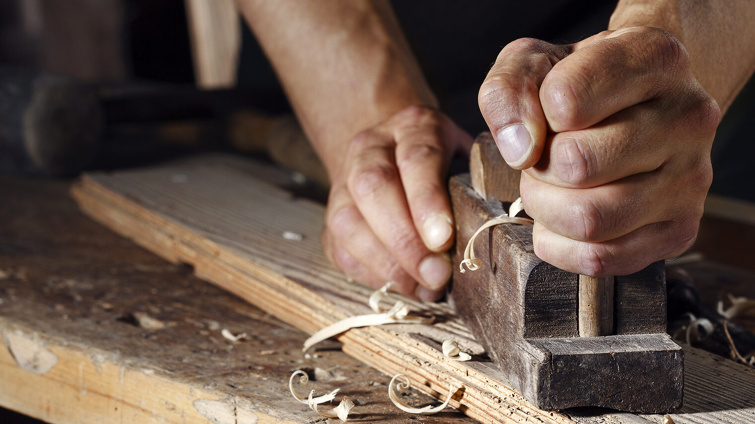 Close up of a carpenter planing a plank of wood with a hand plane Schlagwort(e): adjust, blade, board, building, carpenter, carpentry, chip, closeup, construction, craft, craftsman, craftsmanship, fix, fixing, furniture, hand, handle, hard, hobbies, improvement, joinery, lumber, man, manual, old, plane, planer, planing, plank, professional, repair, sawdust, shaping, sharp, shaving, skilled, skillful, timber, tool, traditional, tree, vintage, wood, wooden, woodwork, woodworking, work, workbench, worker, workshop