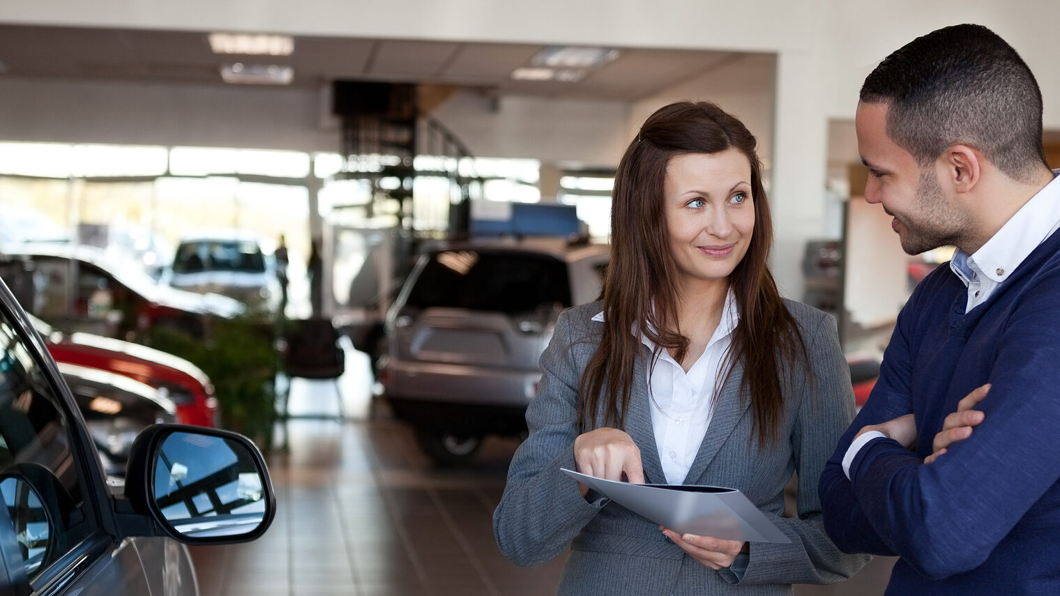 Woman explaining something to a man in a dealership Schlagwort(e): 20s, Young Adult, Man, Male, Mixed-Race, Woman, Female, Caucasian, Indoors, Businesswoman, Suit, Well Dressed, Smart, Elegant, Classy, Stylish, Sophisticated, Communication, Explaining, Interaction, Happy, Smiling, Dealership, Seller, Car, Dealer, Renter, Renting, Garage, Buyer, Vehicle, Automotive, Consumer, Client, Customer, Folder, File, Brunette, Brown Hair, Brunet, Standing, Holding, Showing, Clipboard