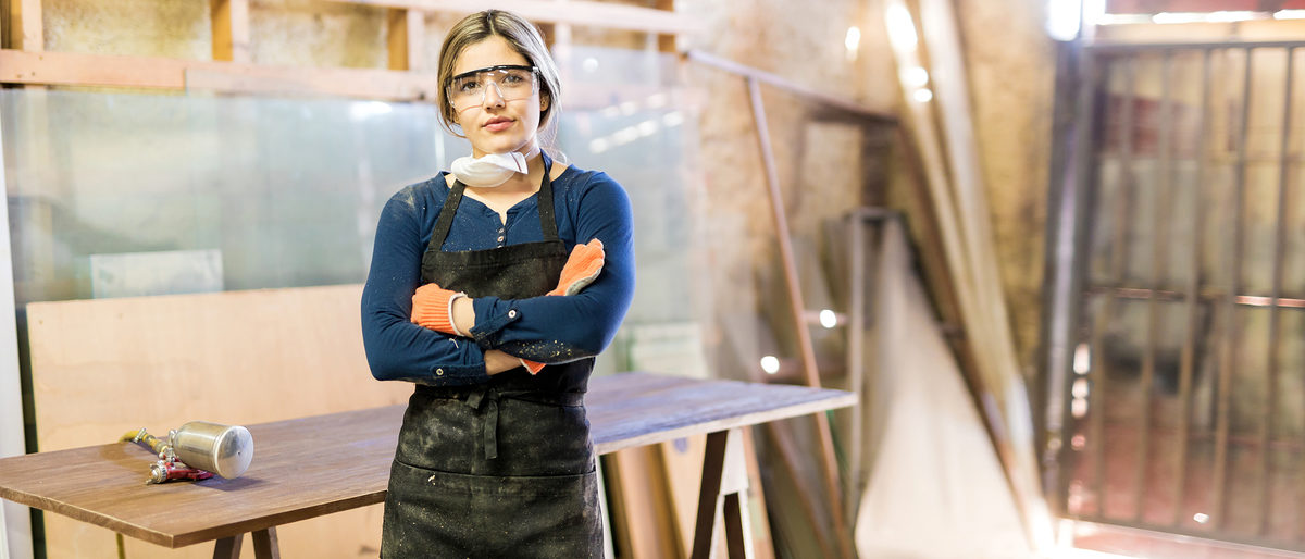 Good looking young confident woman working as carpenter in her own woodshop Schlagwort(e): working, young adult, 20s, equality, gender equality, feminism, carpentry, craft, industry, Latin, Hispanic, female, woman, cute, pretty, beautiful, workshop, woodshop, wood shop, indoor, people, lifestyle, wood, equipment, tool, machine, furniture, worker, carpenter, manual, job, occupation, copy space, copyspace, glasses, protective, gloves, mask, table, varnish, pressure, paint, spray, gun, paint gun, spray gun, arms crossed, eye contact, portrait, woodworking