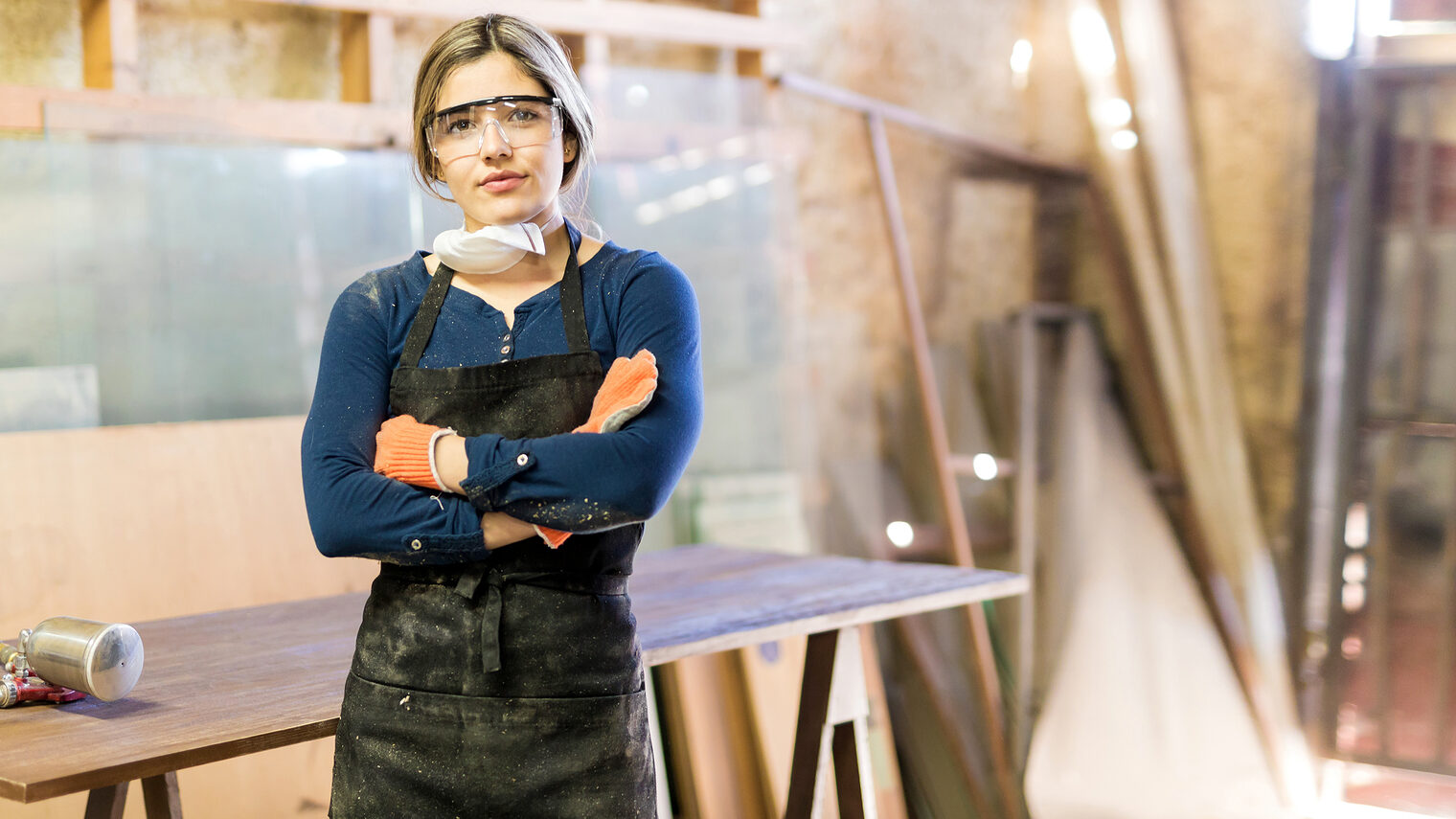 Good looking young confident woman working as carpenter in her own woodshop Schlagwort(e): working, young adult, 20s, equality, gender equality, feminism, carpentry, craft, industry, Latin, Hispanic, female, woman, cute, pretty, beautiful, workshop, woodshop, wood shop, indoor, people, lifestyle, wood, equipment, tool, machine, furniture, worker, carpenter, manual, job, occupation, copy space, copyspace, glasses, protective, gloves, mask, table, varnish, pressure, paint, spray, gun, paint gun, spray gun, arms crossed, eye contact, portrait, woodworking