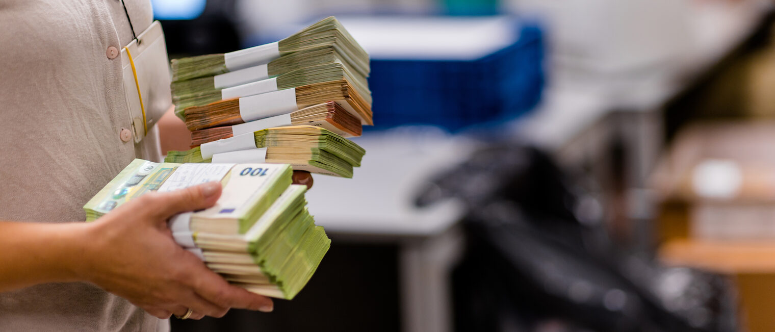 Frau in einer Bank mit Geldbündeln aus Euro-Scheinen. Bild: Dimco / stock.adobe.com