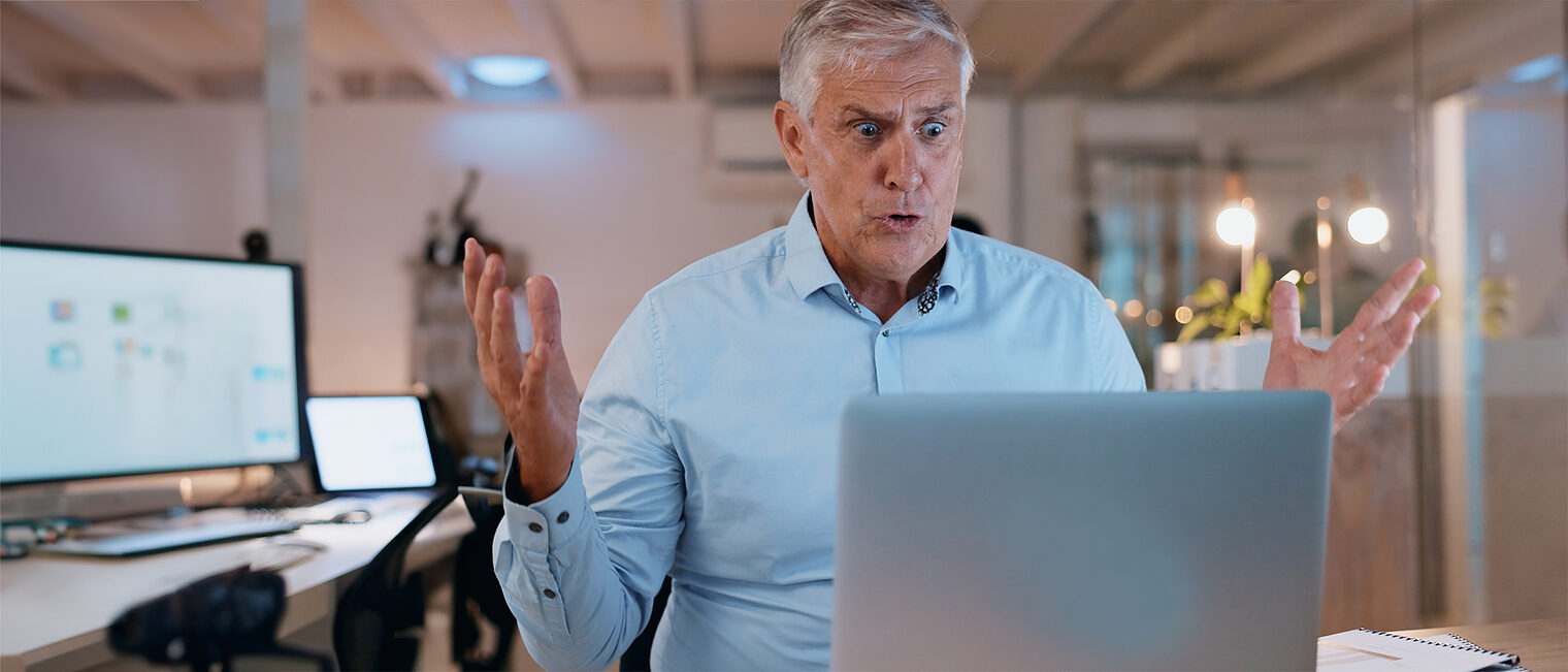 Ein schockierter Unternehmer mit grauem Haar sitzt im Büro vor einem Notebook. Bild: stock.adobe.com / Charlize-D / peopleimages (bearbeitet mit KI / Adobe Firefly)