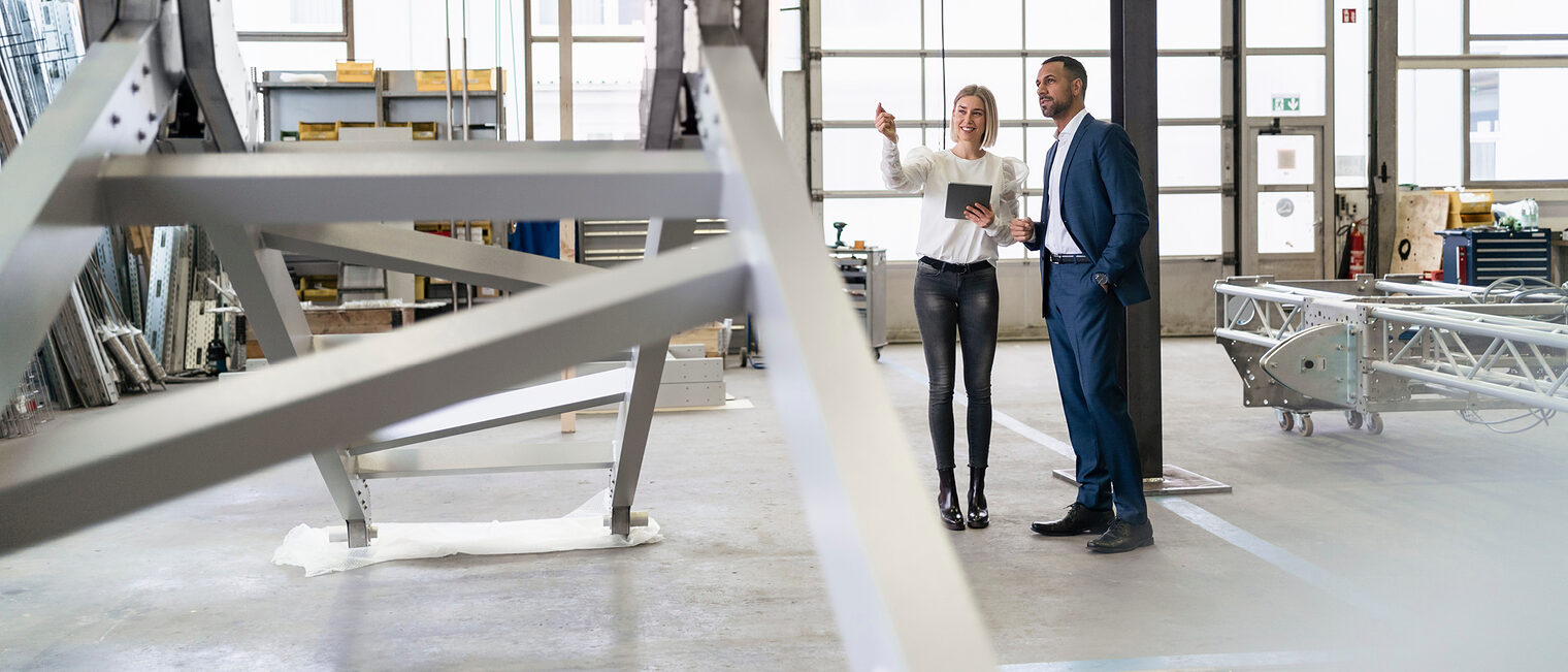 Beratung in einer Werkshalle mit einer Metallbaukonstruktion im Vordergrund. Bild: stock.adobe.com / tunedin 
