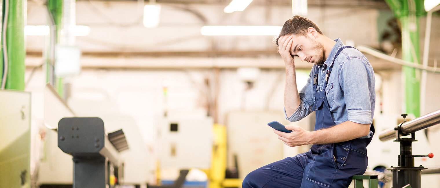 Ein Handwerker sitzt mit einem Smartphone in der Hand sehr nachdenklich in seiner Werkstatt. 