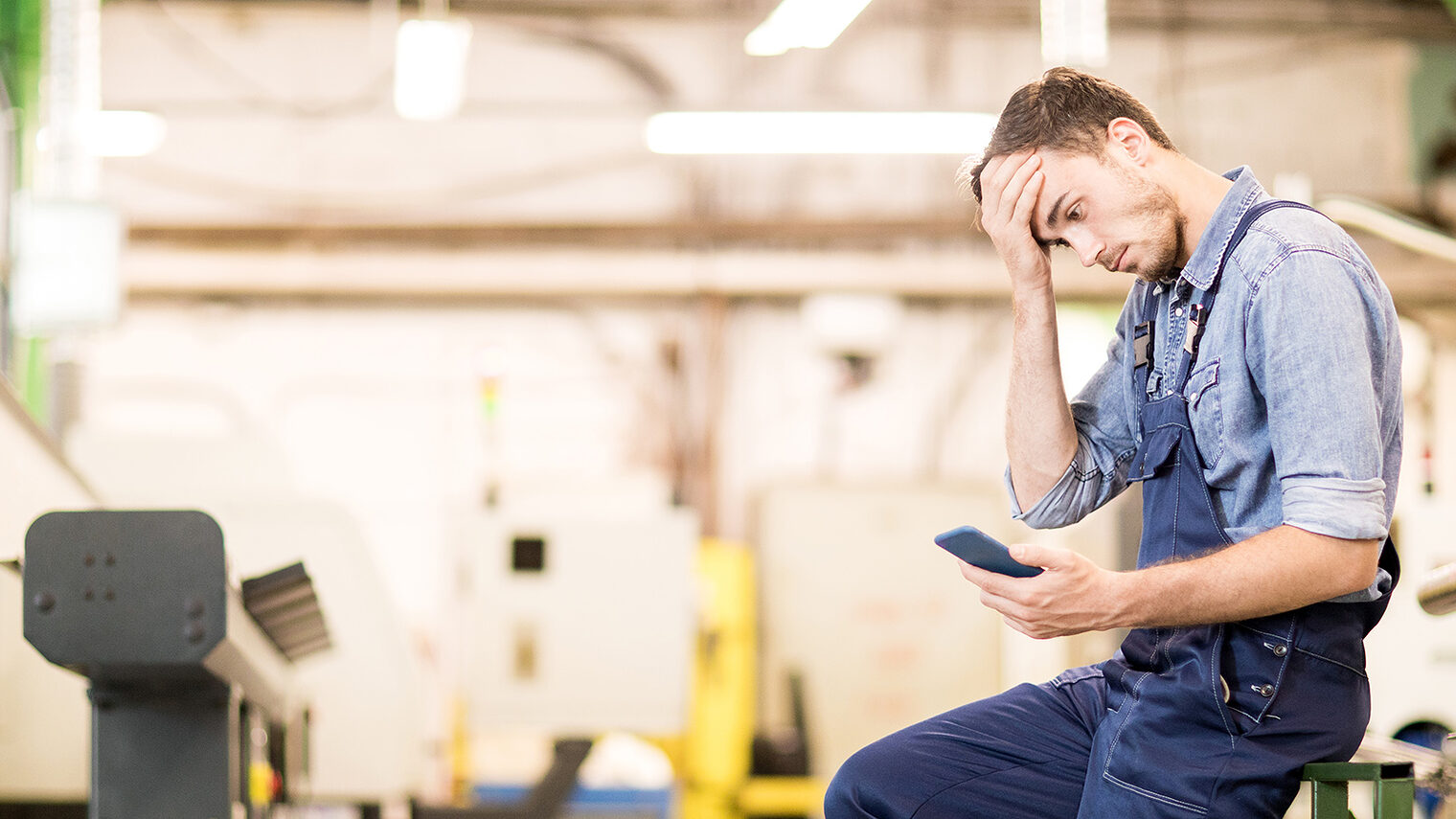 Ein Handwerker sitzt mit einem Smartphone in der Hand sehr nachdenklich in seiner Werkstatt. 