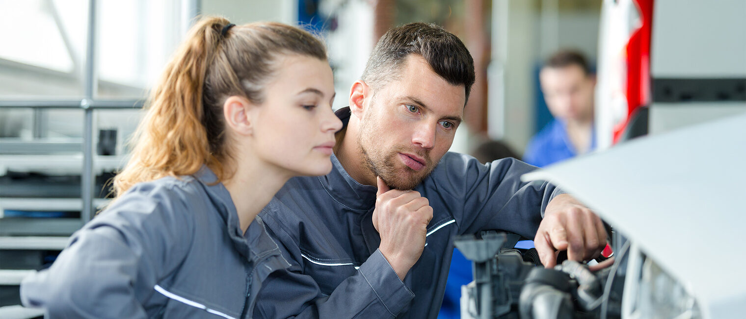 Ein Ausbilder erklärt einem weiblichen Lehrling die Reparatur an einem Auto.