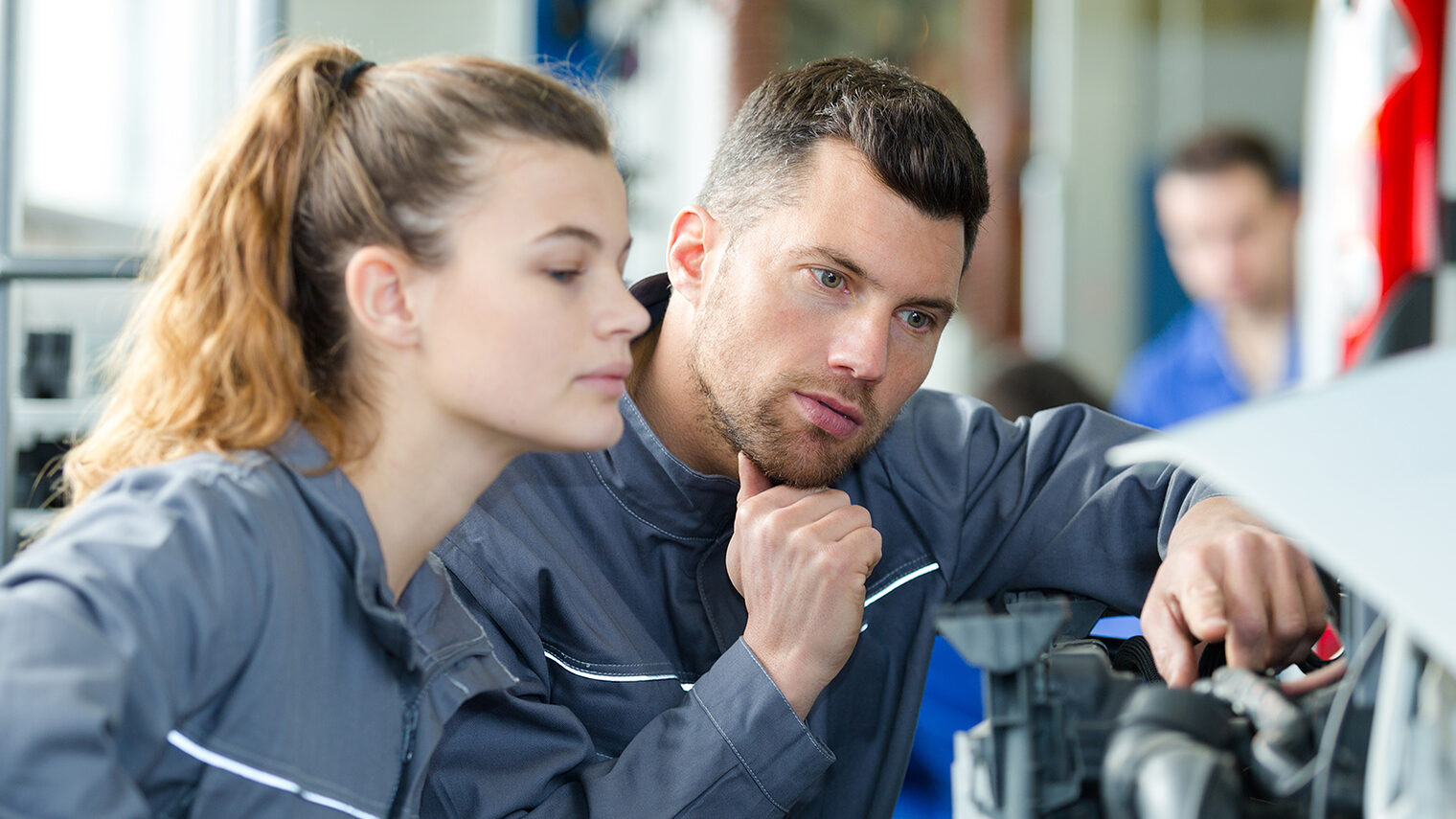Ein Ausbilder erklärt einem weiblichen Lehrling die Reparatur an einem Auto.