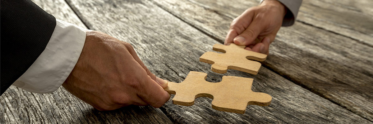 Business partnership or teamwork concept with a business people presenting a matching puzzle piece as they cooperate on finding an answer and solution, close up of their hands. Schlagwort(e): accomplishment, success, teamwork, strategy, solution, business, team, challenge, cooperation, problem, connection, partnership, support, unity, puzzle, pieces, metaphoric, education, assemble, decision, harmony, alliance, coalition, vision, implementation, cooperate, jigsaw, together, businessman, achievement, reunited, relationship, leadership, combination, synergy, concept, development, conceptual, oak, company, two, male, hands, community, collaboration, partners, rustic, wooden, textured, background