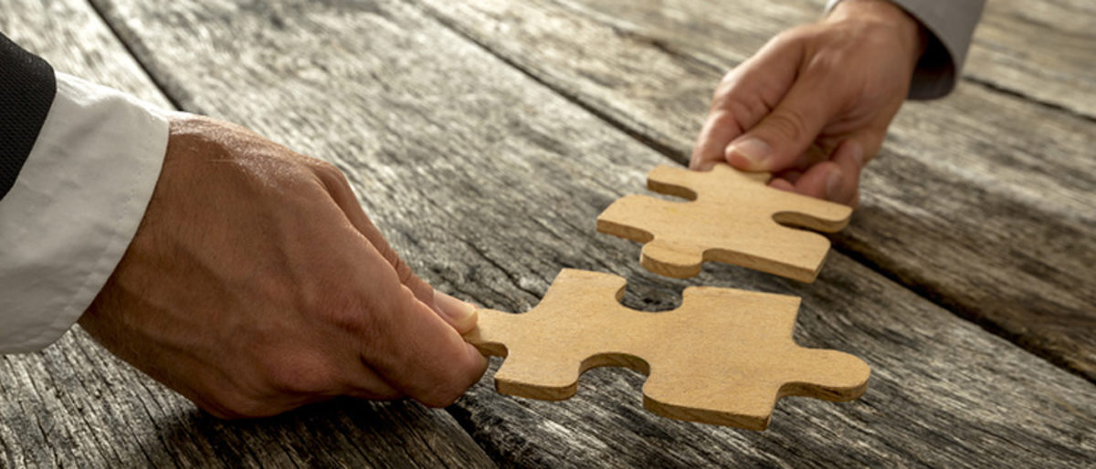 Business partnership or teamwork concept with a business people presenting a matching puzzle piece as they cooperate on finding an answer and solution, close up of their hands. Schlagwort(e): accomplishment, success, teamwork, strategy, solution, business, team, challenge, cooperation, problem, connection, partnership, support, unity, puzzle, pieces, metaphoric, education, assemble, decision, harmony, alliance, coalition, vision, implementation, cooperate, jigsaw, together, businessman, achievement, reunited, relationship, leadership, combination, synergy, concept, development, conceptual, oak, company, two, male, hands, community, collaboration, partners, rustic, wooden, textured, background