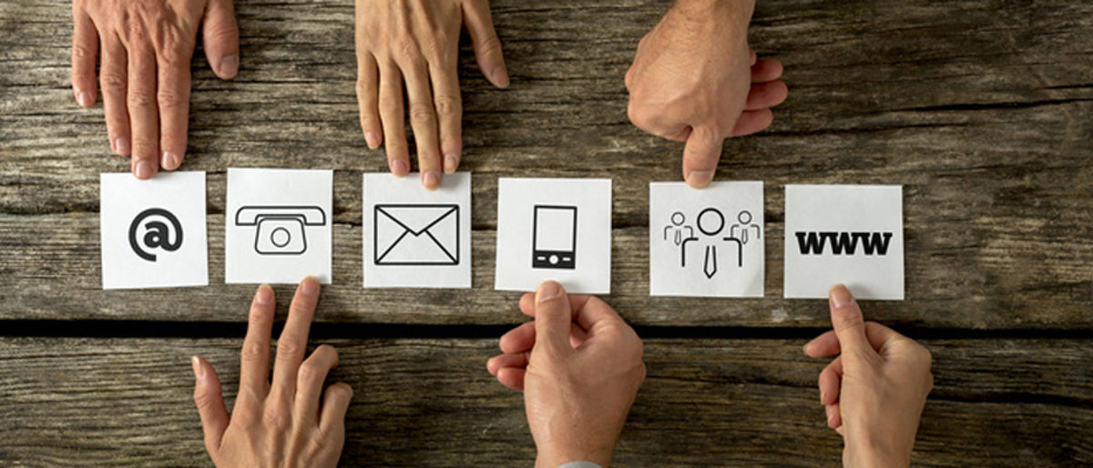 Top view of six people placing white cards with various contact icons in a row on a textured wooden desk. Schlagwort(e): contact, support, customer, service, info, information, business, icon, card, at, sign, phone, telephone, mobile, mail, envelope, email, www, website, businessman, team, company, assistance, helpdesk, businesswoman, 6, six, people, network, helpline, connection, top, overhead, view, hands, male, female, concept