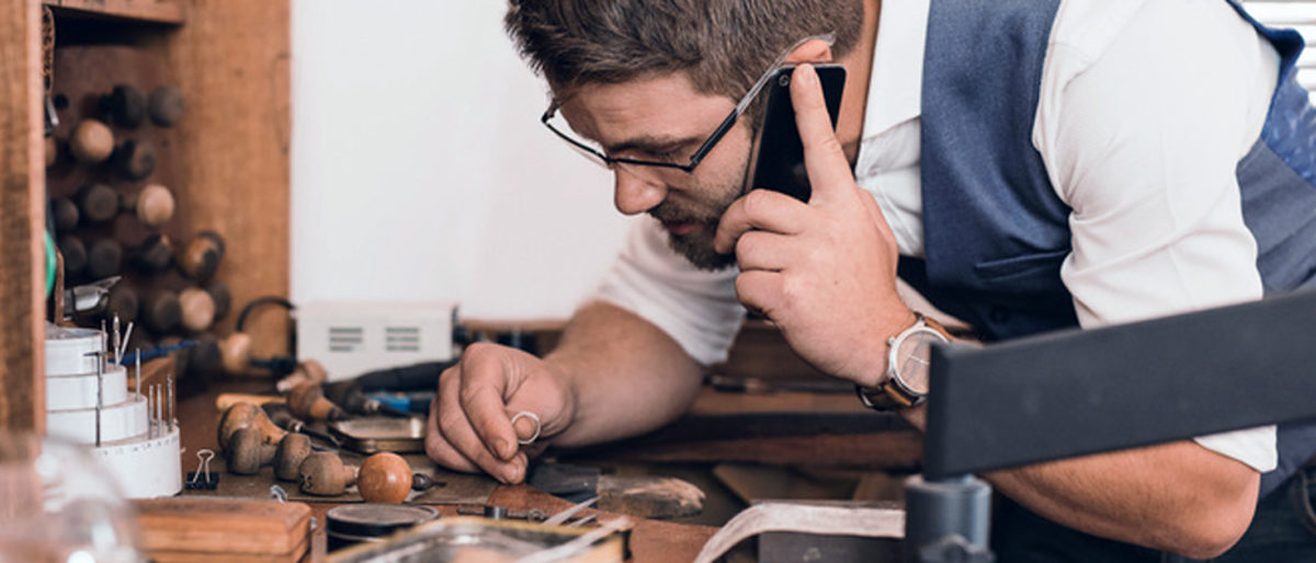 Young jeweler examining a ring and talking on a cellphone while leaning over a workbench full of tools in his shop Schlagwort(e): adult, artisan, caucasian, cellphone, communication, concentration, copy space, craft, crafting, craftsmanship, creative, designer, entrepreneur, equipment, examining, expertise, fixing, focus, glasses, handicraft, handiwork, handmade, holding, industry, inscription, inside, jeweler, jewelry, jewelry design, job, looking, making, male, manufacturing, men, occupation, one, person, phone, profession, reading, skilled, small business, talking, tools, workbench, working, workmanship, workshop, young