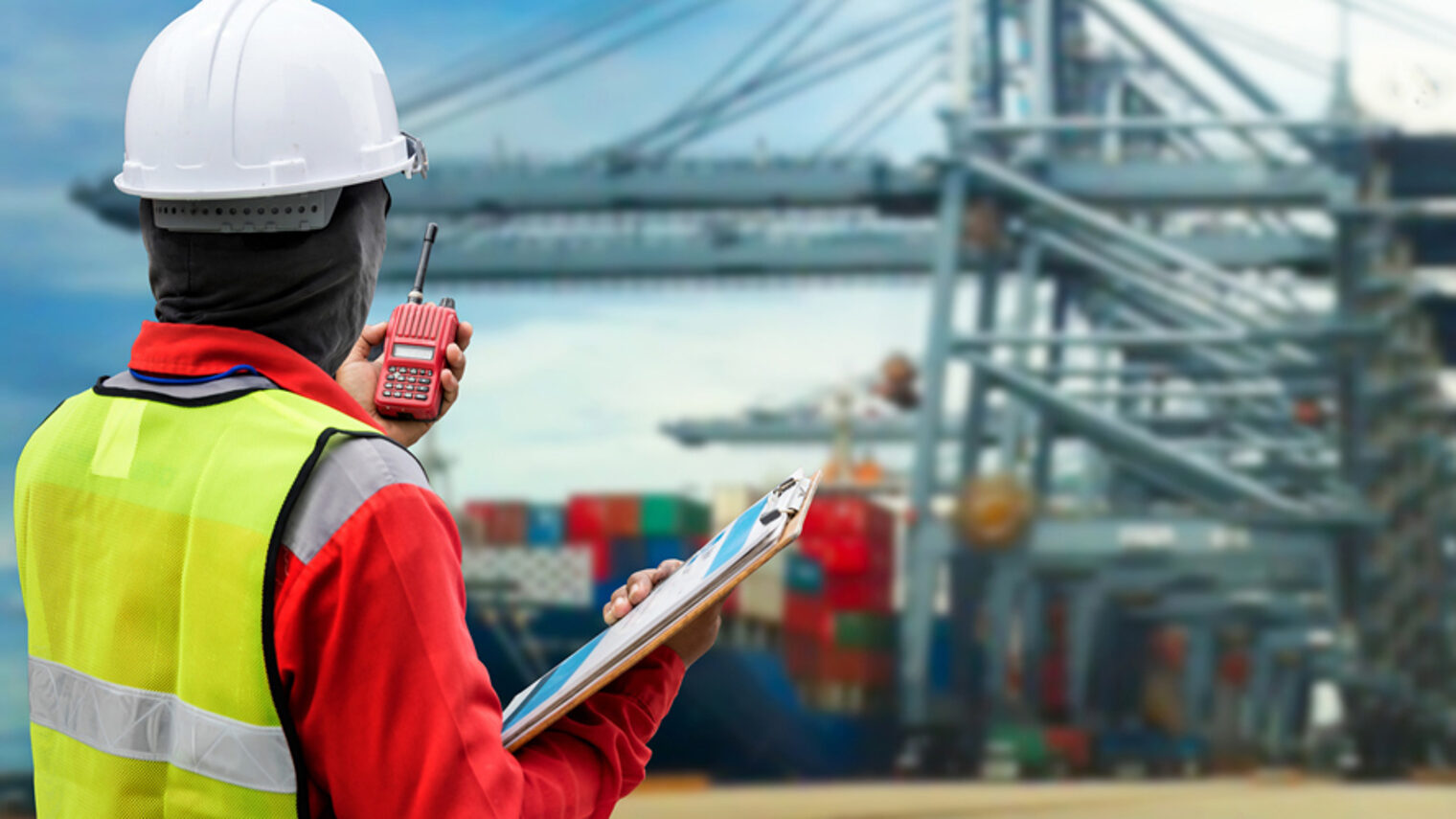 Conceptual image of international logistics, featuring a docker, looking at the unloading of a container ship by huge cranes in the Schlagwort(e): transport, transportation, freight, concept, shipping, port, export, bulk, docker, import, international, modern, huge, goods, technology, hard hat, conceptual, large, logistics, overalls, multinational, economics, economy, coveralls, red, looking at, industrial, safety, containers, commerce, industry, container ship, harbor