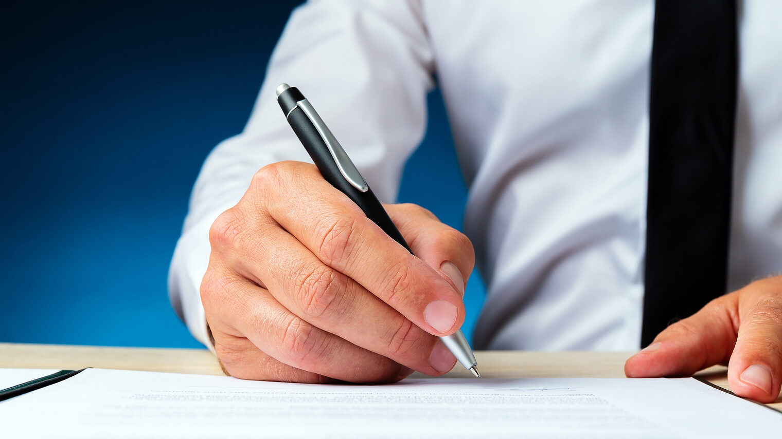 Business manager signing a document in a folder on his desk. Over dark blue background. Schlagwort(e): document, contract, legal, signing, form, deal, business, businessman, manager, lawyer, insurance, pen, successful, paperwork, signature, executive, agreement, write, official, employment, broker, occupation, formal, note, documentation, businessperson, subscription, lease, file, loan, plan, report, testament, attorney, employer, final, statement, realtor, briefing, approved, investor, economy, folder, corporate, working, entrepreneur, career, blue, background